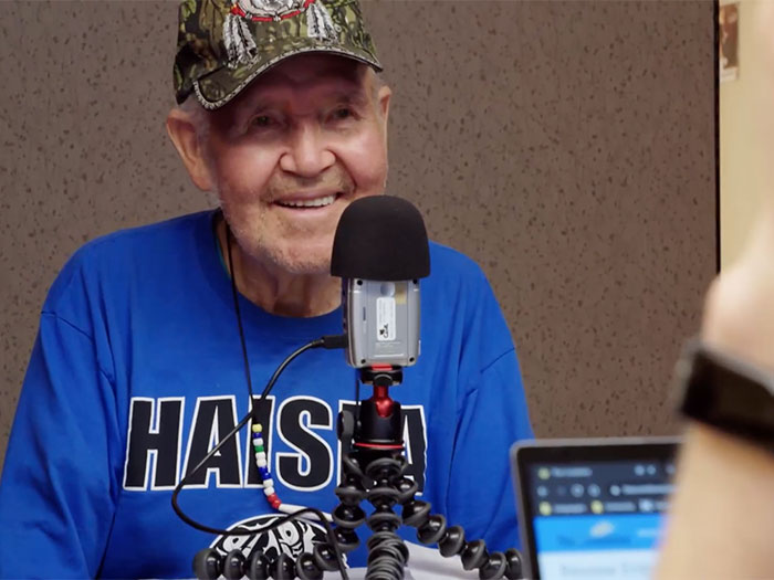 An elderly man wearing a camo baseball cap and blue sweatshirt smiles while speaking into a microphone. He appears to be participating in a podcast or radio interview, with a laptop visible in the foreground.