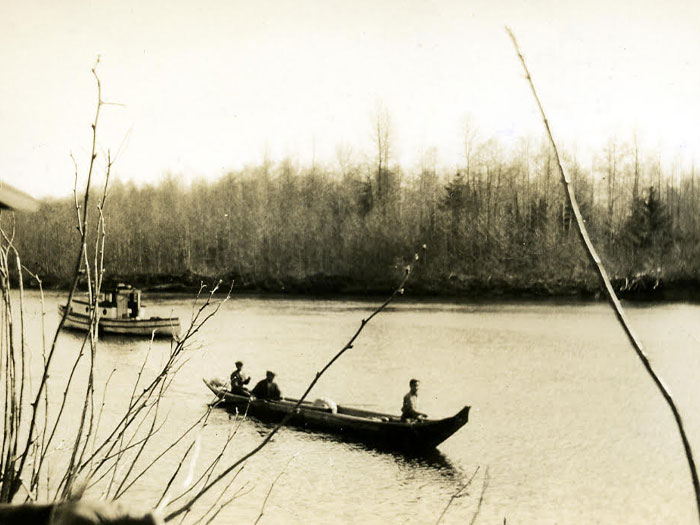 Three people are visible in a long wooden boat on a calm river, with a larger boat in the background. Bare, leafless trees line the riverbank under an overcast sky, creating a serene, late autumn or early spring scene.