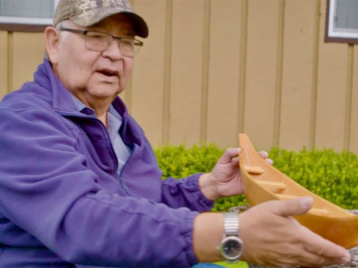 An elderly person wearing a camo cap and a purple jacket holds a small wooden canoe model. They are seated outdoors in front of a wooden building with greenery in the background. The person appears to be discussing or examining the model.